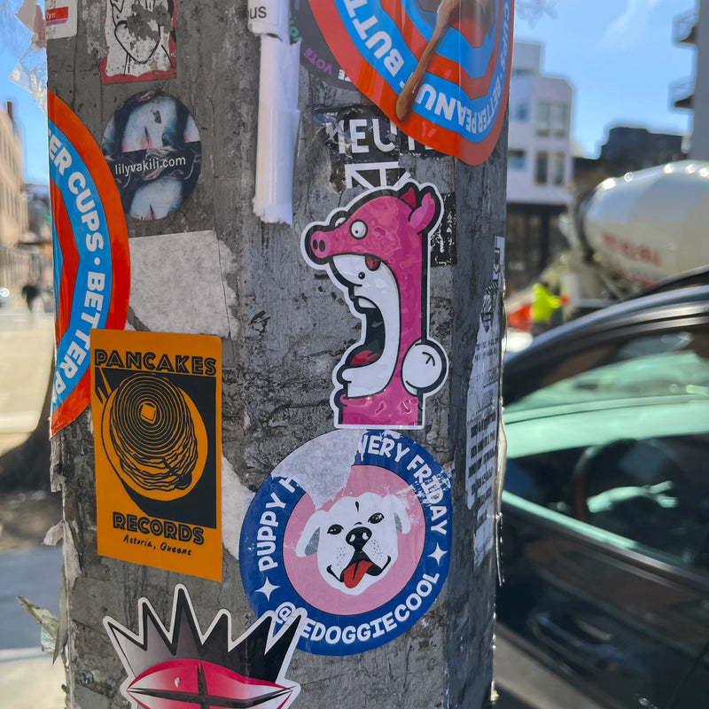 A close-up of a concrete utility pole covered with multiple colorful stickers, including a Shouter vs Pig sticker by Ran Kunst